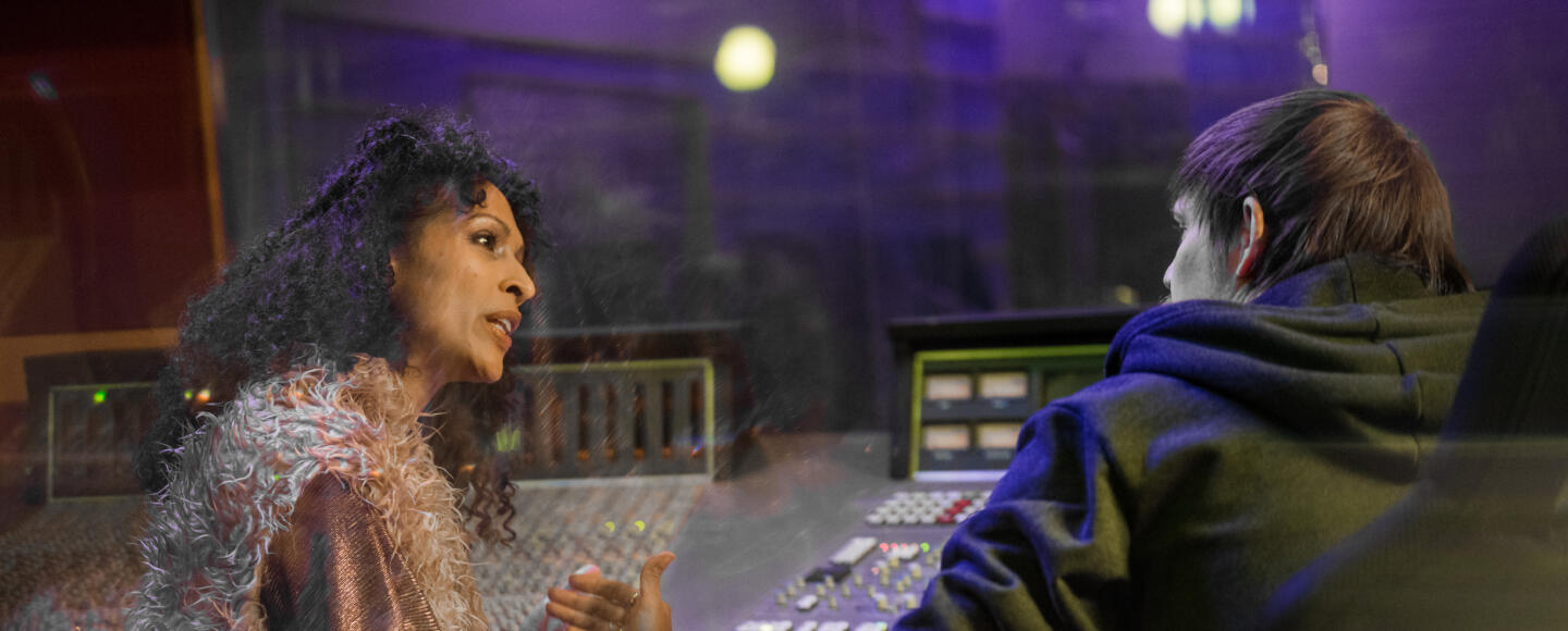 A woman and a man are sat together at a sound desk. The woman appears to be offering advice to him.