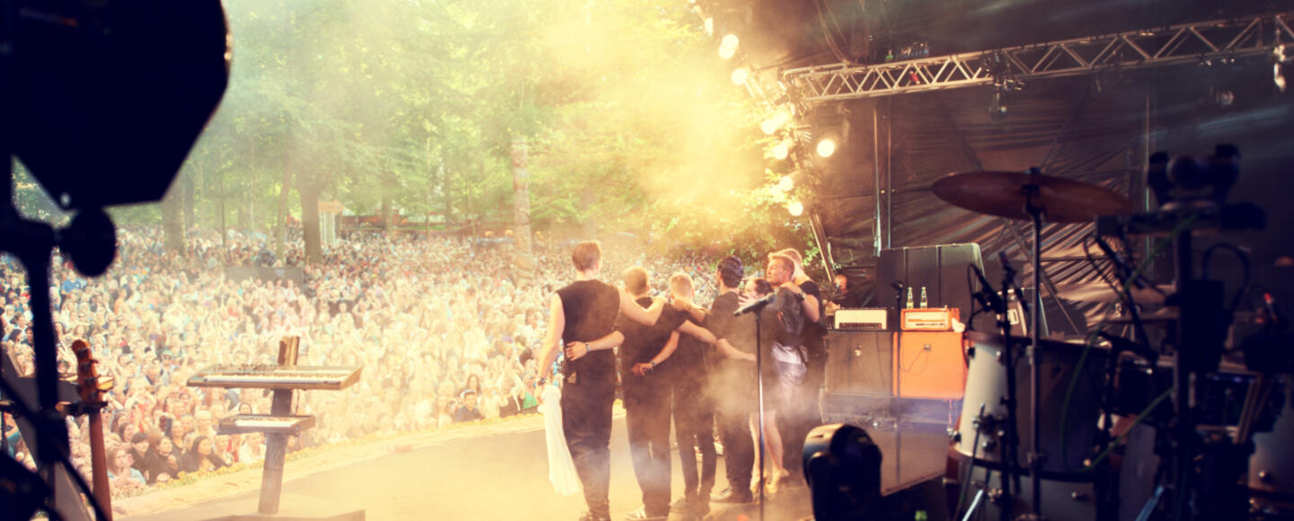 A band lock arms onstage facing a large outdoor audience